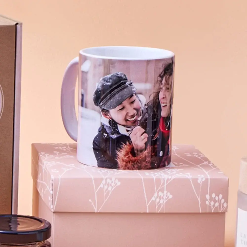 Two personalised mugs on a wooden table, one decorated with festive gingerbread and candy cane designs featuring the name Eloise, accompanied by a small plate of cookies, with a blurred Christmas tree in the background.
